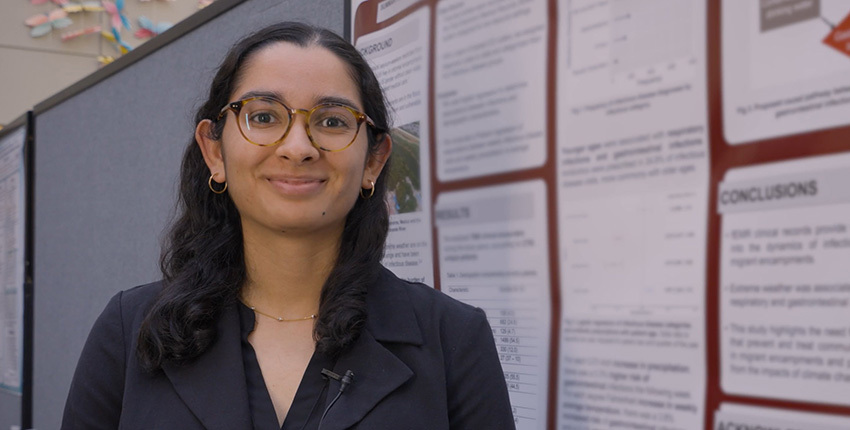 A young woman wearing a dark blazer and wearing glasses stands smiling beside research poster.