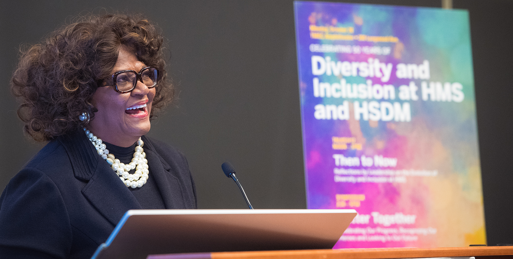 Alt Text:  A woman standing at a lectern, speaking in front of a colorful, out-of-focus poster