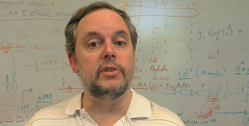 White man with beard stands in front of a whiteboard covered in colorful schematics