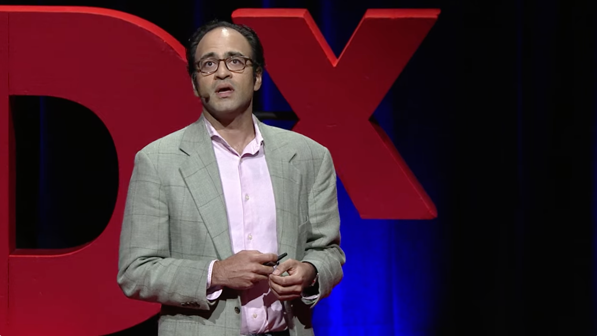 Screen shot of a South Asian man in a suit mid-sentence in front of the TEDx logo on stage.