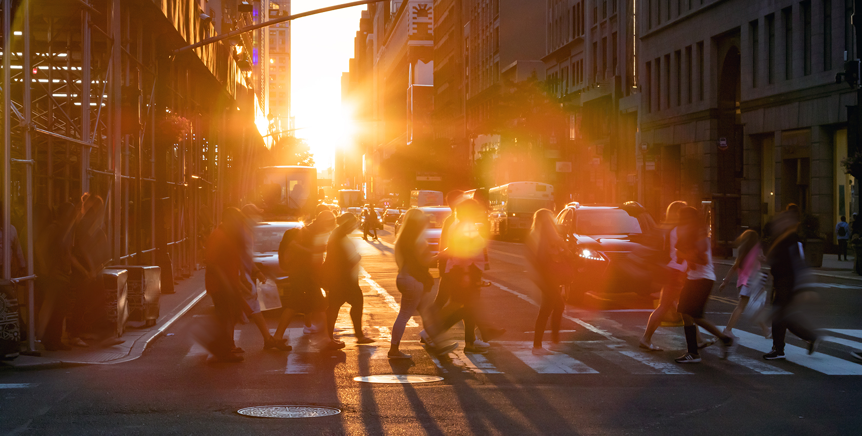 A group of people cross a street at a crosswalk, backlit by the glow of the sun and surrounded by a halo of rainbowed lens flare.