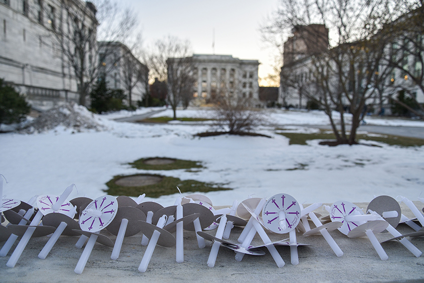 Unlit candles and paper holders line a stone wall at the edge of a snowy HMS Quadrangle, with Gordon Hall visible in background