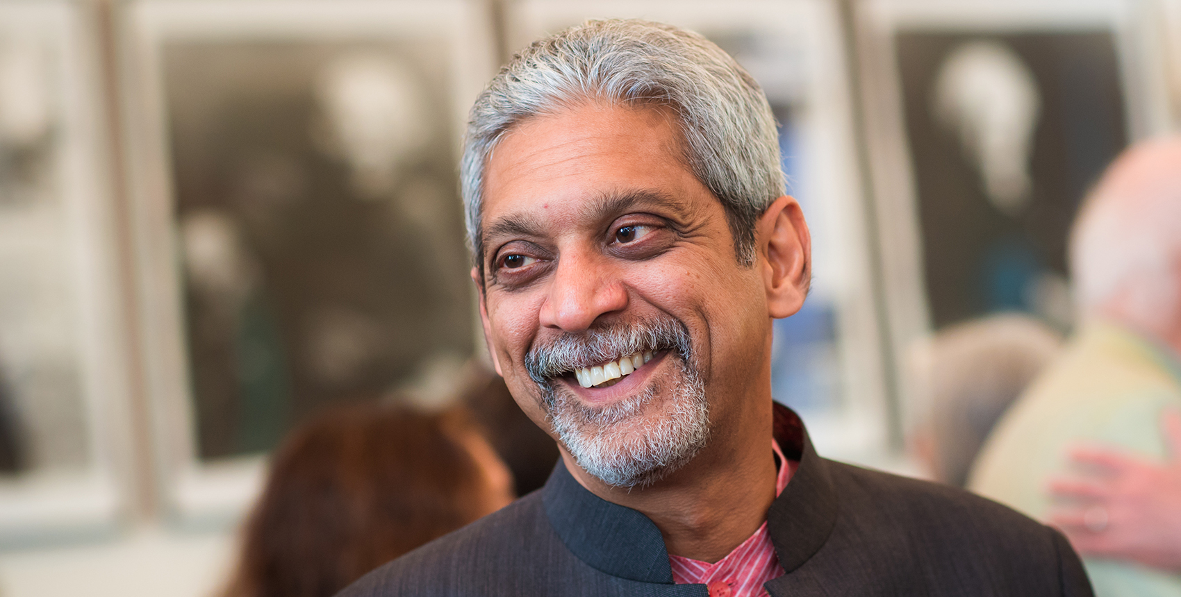 A silver-haired man, wearing a black jacket and a red shirt, smiles at someone off camera.