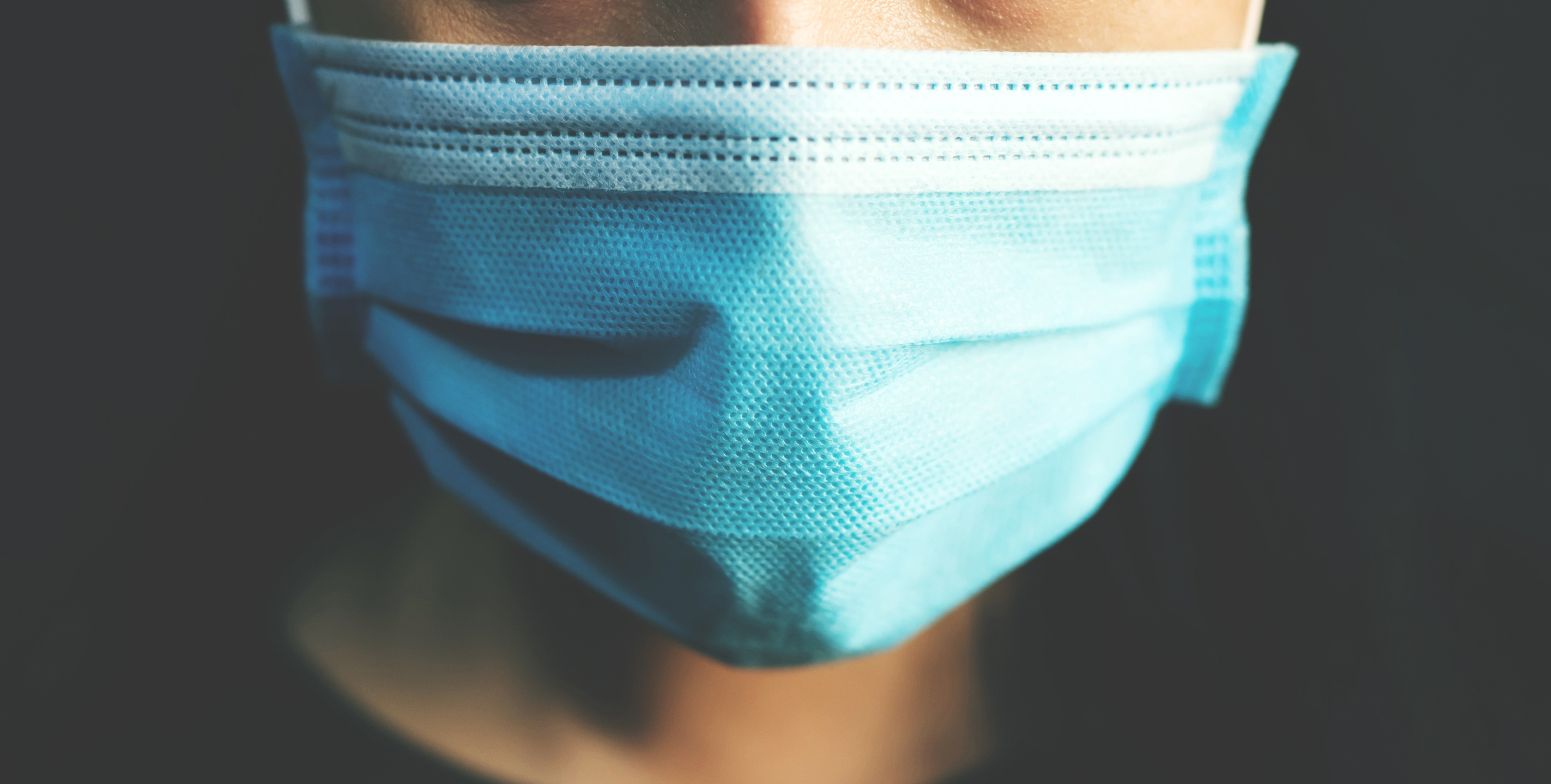 Closeup of the face of a person wearing a paper medical mask in front of a dark background.