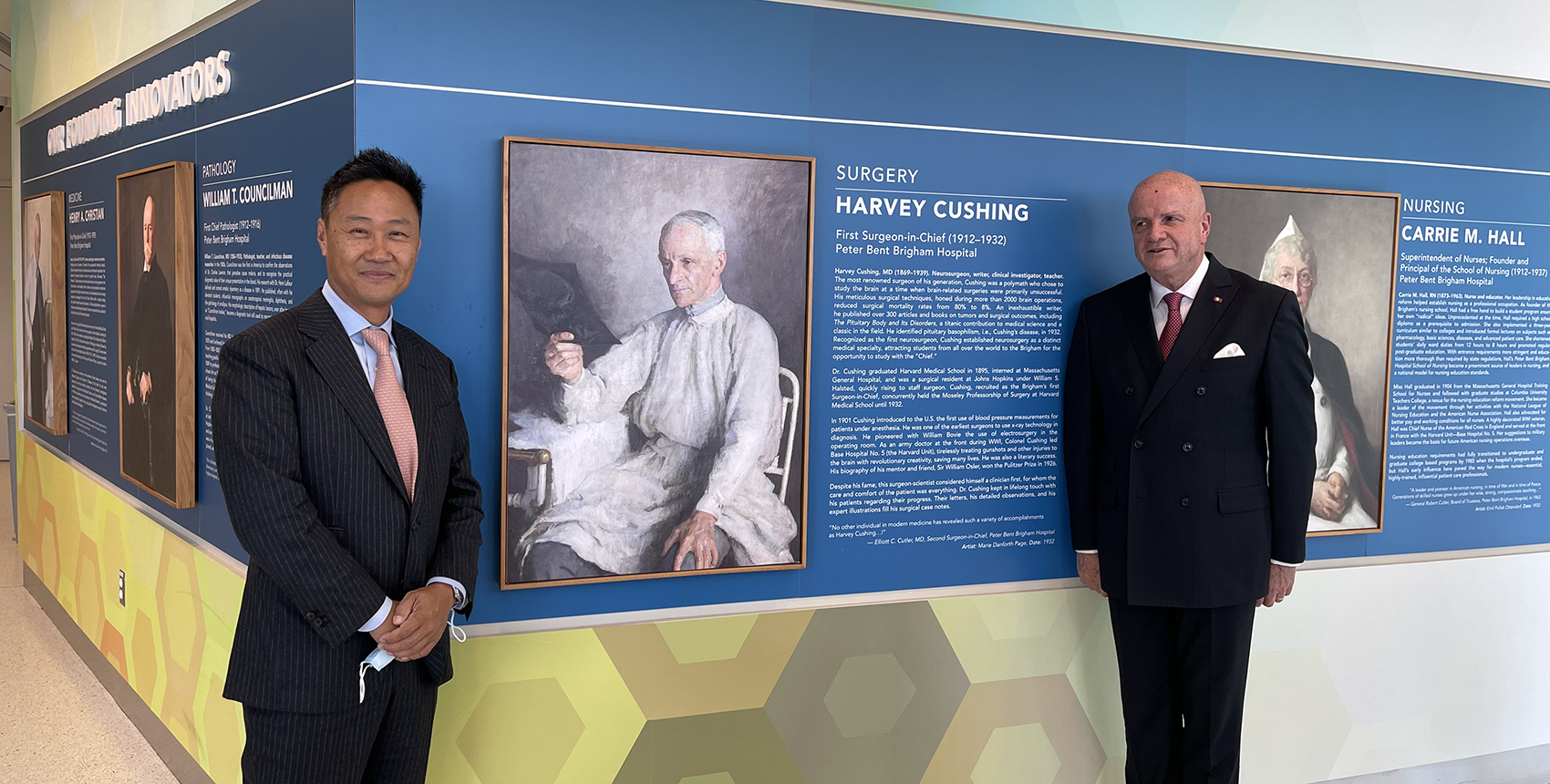 In a hallway painted with colorful geometric patterns, two men in suits stand on either side of a portrait of a man in surgical gowns reviewing an X-ray of a patient’s skull. The portrait is labeled “Harvey Cushing.”