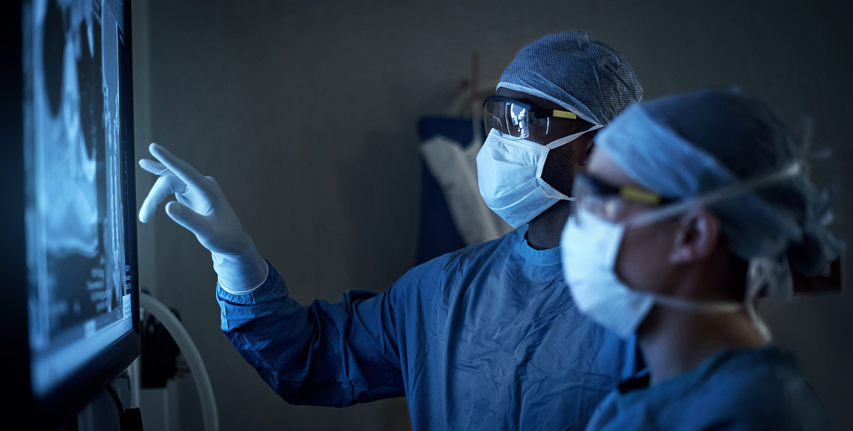 Two figures wearing surgical masks, gowns, and caps examine a backlit X-ray image