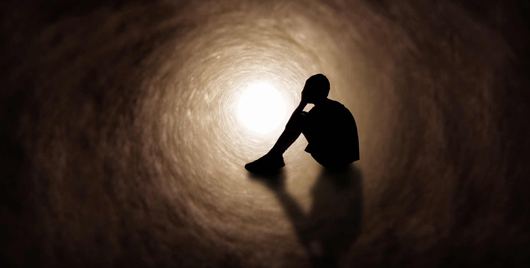 The silhouette of a young person in shorts and t-shirt covering their face as they sit in a dark tunnel.