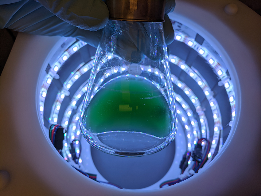 A flask of green liquid held by a gloved hand is silhouetted at the top of a cylinder with rings of lights