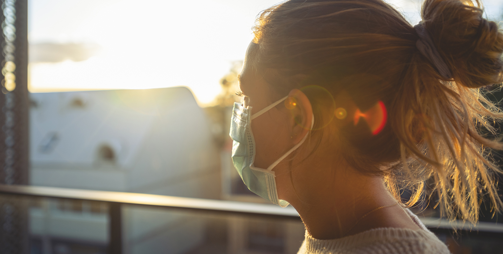 A woman  wearing a mask is seen in profile, backlit by the sun.