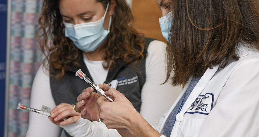 Two women in white coats and surgical masks hold syringes labeled "COVID-19 vaccine"