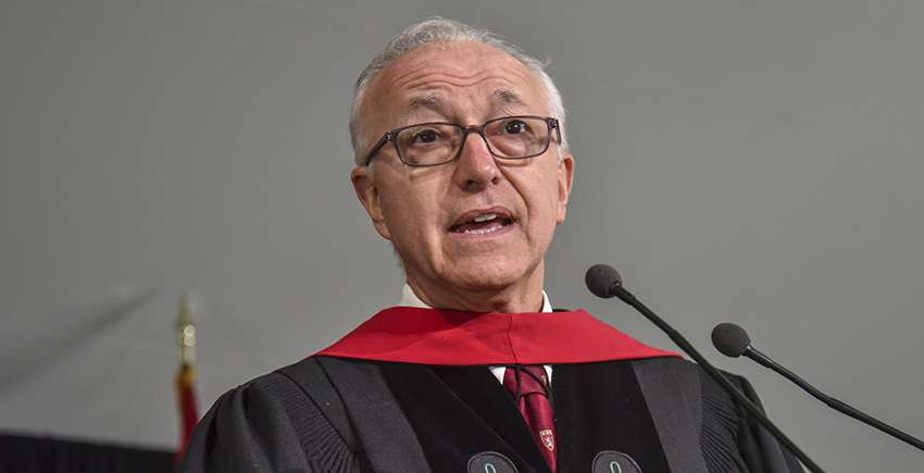 A gray-haired man with glasses and doctoral robes speaks into a microphone