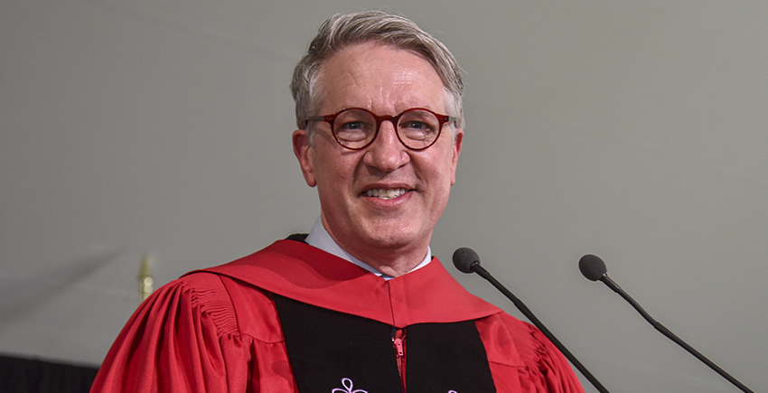 A white man in glasses and red robes smiles at a microphone