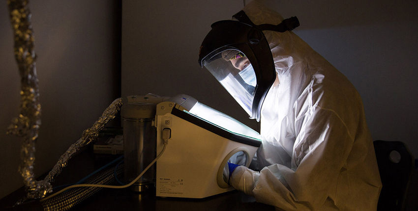 Person in a full-body clean suit and masked hood leans over a lit display in a dark room