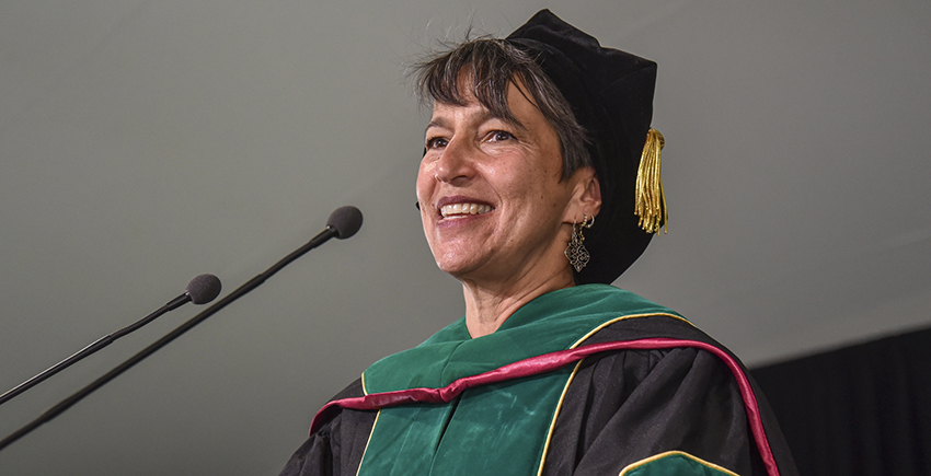 Close-up of a white woman with gray hair in doctoral robes smiling as she speaks into a microphone
