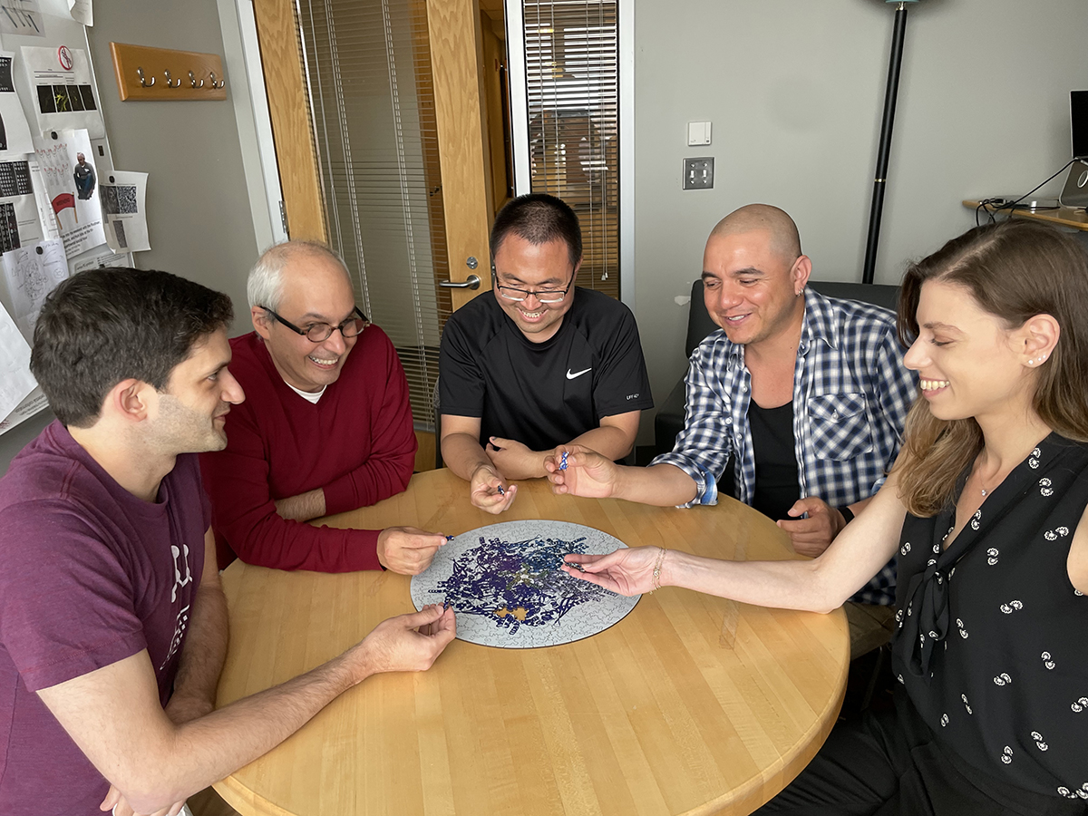 Five people sitting around a wooden table with a jigsaw puzzle in the middle of the table.
