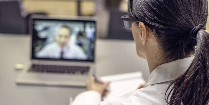 Looking over a doctor's shoulder, we see a patient video conferencing on the doctor's laptop.