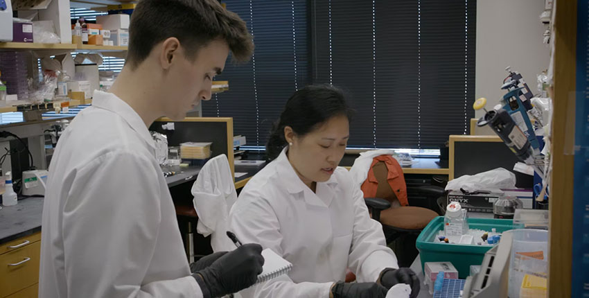 Two researchers in lab coats work at a bench. One is taking notes.