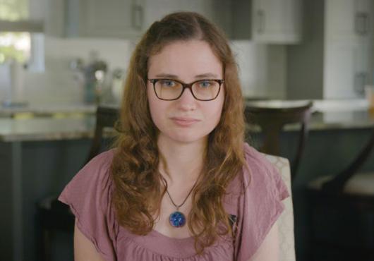 A shot of Jessica Chaikof from the chest up, wearing a pink shirt and glasses
