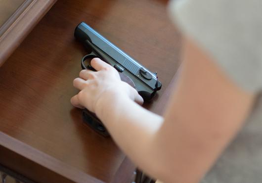 A child's hand reaches into an open drawer in a wooden dresser to pick up a black handgun with a brown handle.