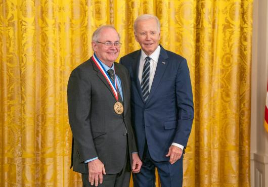 U.S. President Joseph Biden stands in a suit beside Gregory Petsko, a gray-haired white man also in a suit. Petsko wears his new National Medal of Science. Both are smiling. They stand before a gold curtain.
