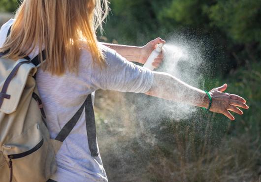 white woman wearing a backpack, viewed from behind, spraying insect repellant onto her bare forearm in a field next to some woods