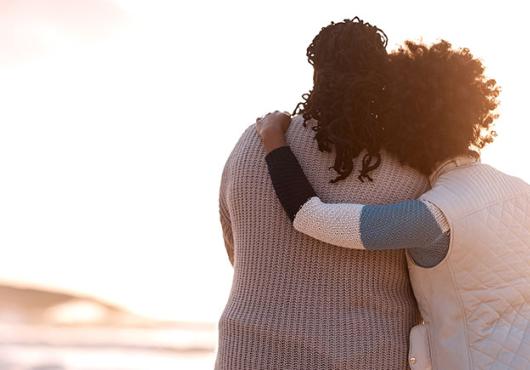 Two women stand side by side embracing one another on a beach facing the ocean.