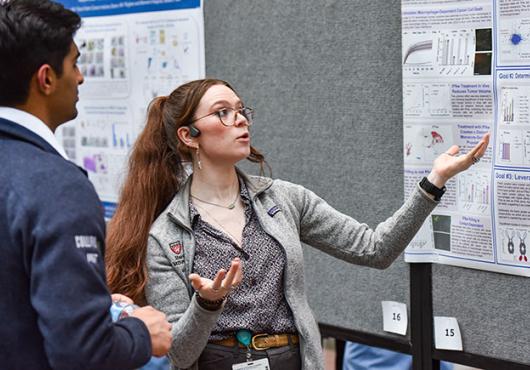 A young woman stands in front of research poster motioning with her hands while a young man listens to her explanation.