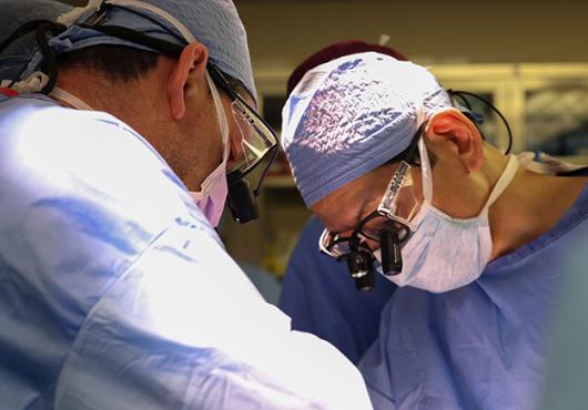 Photo of surgeons in scrubs and masks operating on an unseen patient. Only one is seen head-on. He wears attachments on his glasses to magnify what he's seeing.