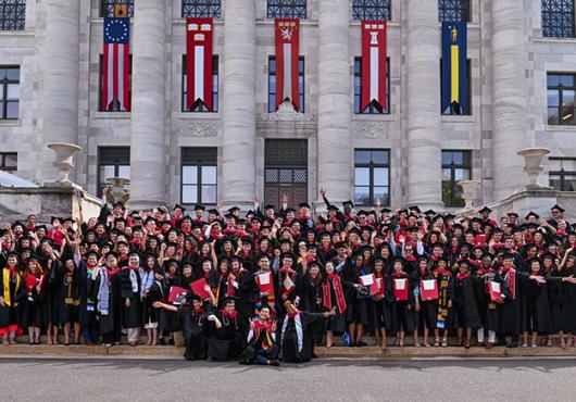 Wide-angle photograph of MD and DMD graduates in regalia cheering on the front steps of Gordon Hall, five tall flags hung between marble columns behind them