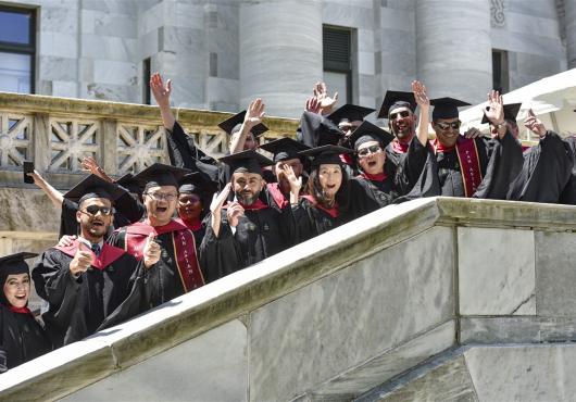 More than a dozen graduates in regalia cheer on the marble stairway at one side of HMS's Gordon Hall