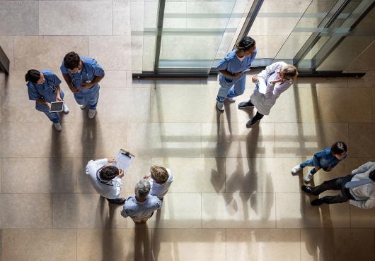 An overhead view of nine people in the entryway of a hospital, standing in groups of two or three, many dressed in medical gear or lab coats, carrying clipboards and digital devices.