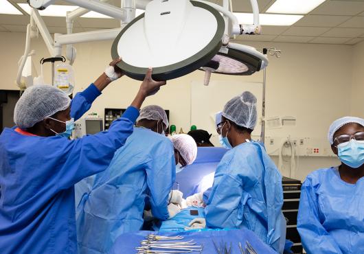 A surgical team of doctors and nurses wearing blue scrubs, gauzy head coverings, and medical masks adjust lights, provide anesthesia, and perform other actions necessary for an operation, all gathered around the patient on the operating table. Health equity requires access to surgical care.