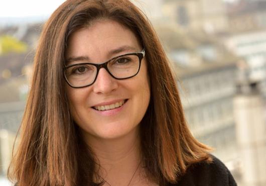 Portrait photo of a smiling woman with glasses and shoulder-length hair, sitting on a wall with a city skyline out of focus in the background.