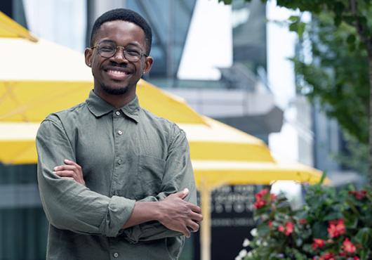 A smiling young Black man with glasses stands with arms crossed in front of picnic tables on the HMS Quad
