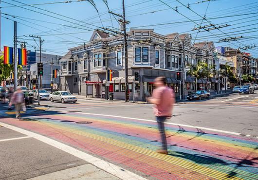 People in colorful outfits cross the street at an intersection with rainbow crosswalks in San Francisco’s Castro district.