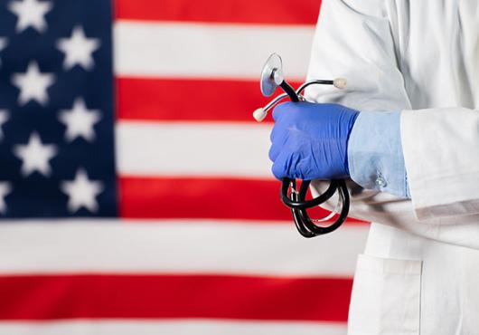 A health care practitioner wearing a white coat and holding a stethoscope stands in front of an American flag