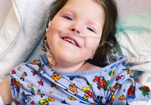 Photo of a young girl lying on blankets wearing a hospital gown featuring cartoon characters.