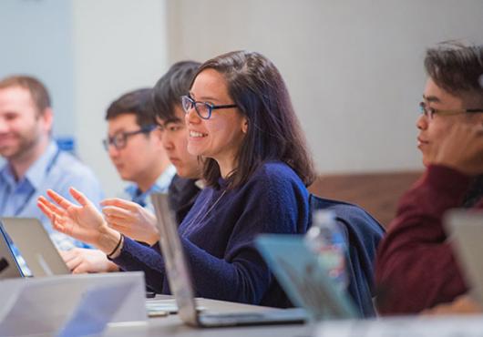 Six trainees at a conference table with laptops in front of them. The one at center speaks with a smile and gesturing hands.
