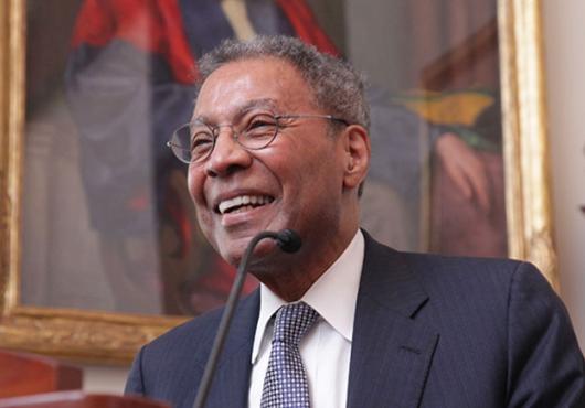 A man in a suit smiles at a lectern in front of a painting of someone in academic robes