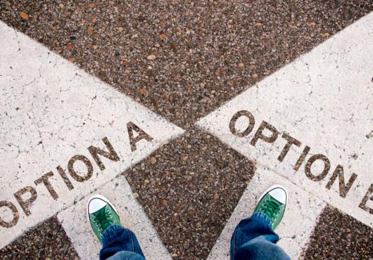 An aerial view of two feet in green sneakers, one standing on a large white arrow labeled “option A” and the other standing on a large white arrow labeled “option B”