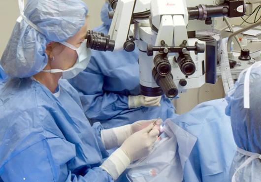 In an operating room, three people in blue surgical gowns, masks, and head covers look through microscopes while performing surgery on a patient.