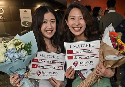 Two smiling women hold bouquets and Match Day signs from Harvard Medical School, celebrating their residency placements in Medicine + Neurology and Dermatology.