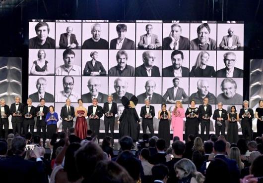 A row of Breakthrough laureates standing on a stage with black and white headshots of previous winners projected in the background