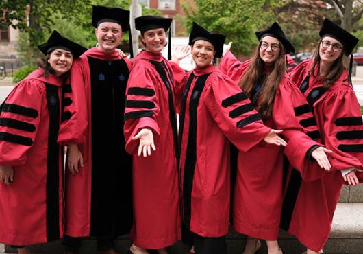 Five women and one man in academic regalia smile and stretch out their hands, tah dah.