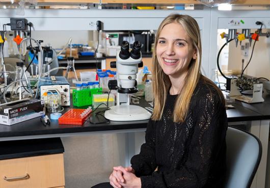 A researcher sits on a chair next to a microscope in a lab space
