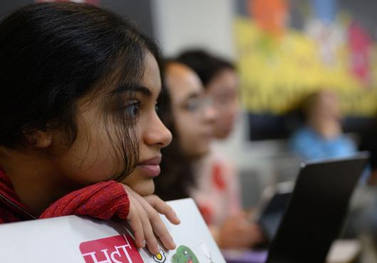 Focus on one student leaning on laptop with an HST sticker on the cover with several students in blurred background.