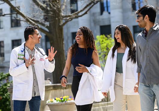 Four medical students, two in white coats, smiling and walking together on a sunny day with a marble columned building in background.
