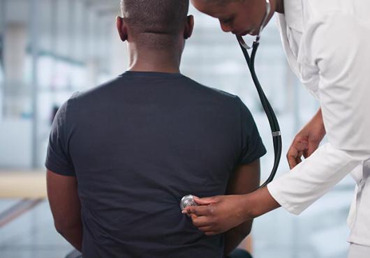 A doctor in a white lab coat uses a stethoscope to listen to the back of a seated patient wearing a dark t-shirt, in a bright, modern medical office.
