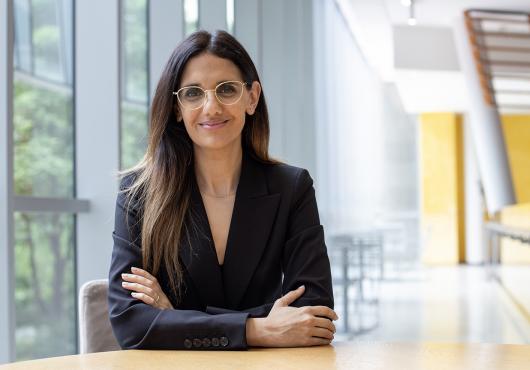 A woman with long, straight hair and glasses sits at a round wooden table in a modern, sunlit building with large windows. She is wearing a black blazer and smiling confidently with arms crossed.