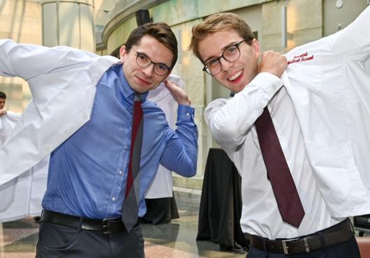 Two men put on white coats in a sun-filled atrium.